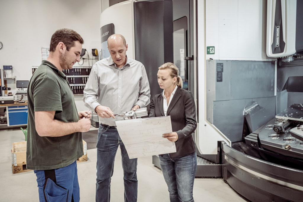 Drei Personen stehen in einer Produktionsstätte und besprechen Pläne. Ein Mann in einem grünen Hemd, ein anderer in einem grauen Hemd und eine Frau im Anzug halten technische Zeichnungen in der Nähe von Industriemaschinen und prüfen sie.