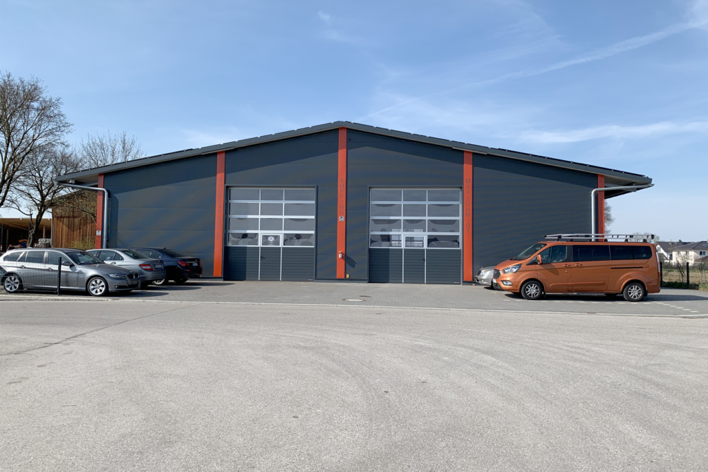 Ein großes, modernes Garagengebäude mit drei Metalltoren, jedes mit Fenstern. Davor parken mehrere Autos, die Außenfassade ist grau mit orangefarbenen Akzenten. Der Himmel ist klar und blau.