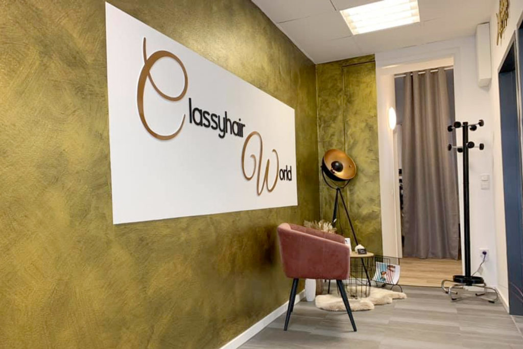 Ein Salon-Interieur mit olivgrünen Wänden und einem „Classyhair World“-Schild. Es gibt einen rosa Stuhl mit weißem Fell, eine Stativlampe und eine Garderobe. Helle Deckenleuchten und ein Flur mit einem Vorhang im Hintergrund runden den Look ab.