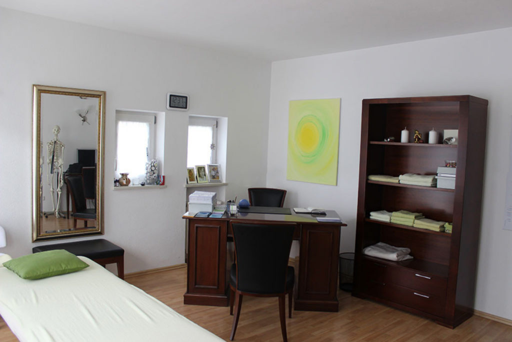 Ein aufgeräumtes Büro mit einem Holzschreibtisch und Stuhl in der Mitte, einem hellgrünen Massagetisch mit einem Kissen auf der linken Seite, einem großen Spiegel an der Wand und einem Bücherregal mit ordentlich gefalteten Handtüchern auf der rechten Seite. An der Wand hängt ein helles Gemälde.