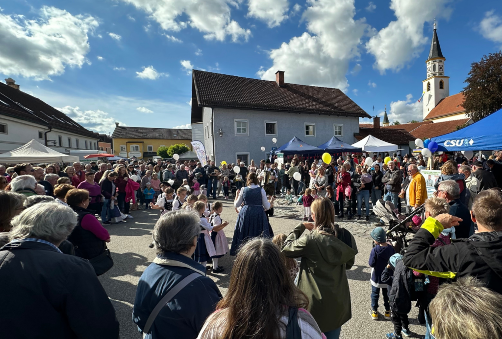 Ein lebhaftes Open-Air-Festival mit Menschen, die sich auf einem Stadtplatz versammeln. Kinder in traditionellen Kostümen treten im Zentrum auf. Die Szene ist sonnig mit blauem Himmel, umgeben von alten Gebäuden und einem Kirchturm, während Zuschauer zuschauen und Fotos machen.