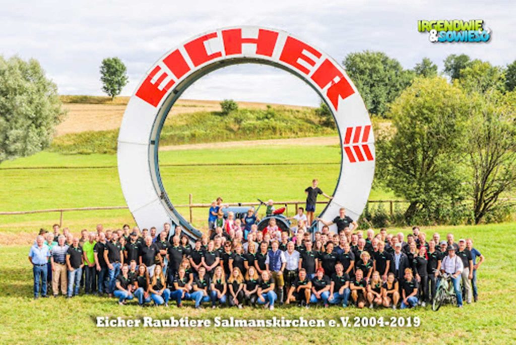 Eine große Gruppe von Menschen posiert vor einem riesigen Eicher-Logo auf einer Wiese mit Bäumen und sanften Hügeln im Hintergrund. Viele tragen Freizeitkleidung und einige fahren Fahrrad. Der Text lautet „Eicher Raubtiere Salmanskirchen e.V. 2004-2019“.