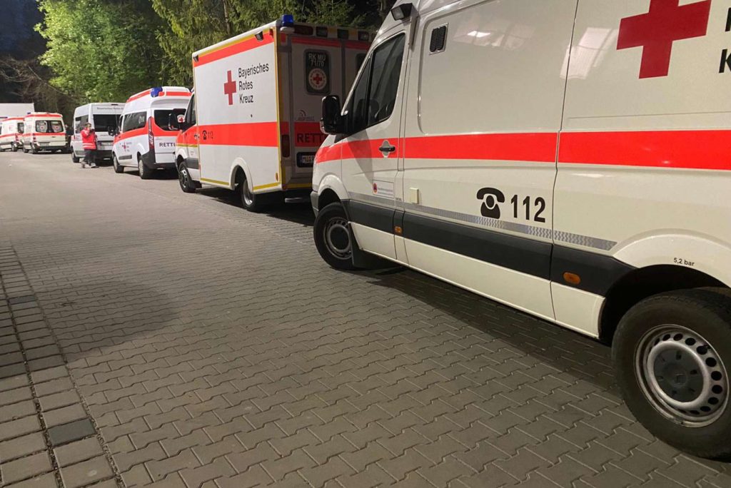 Eine Reihe von Krankenwagen mit rot-weißem Design parkt nachts auf einer gepflasterten Straße. Auf einem Fahrzeug sind „112“ und „Bayerisches Rotes Kreuz“ auf der Seite zu sehen. Im Hintergrund sind Bäume zu sehen.