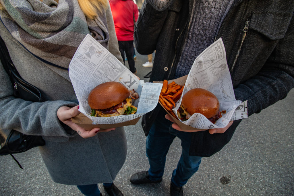 Zwei Personen, eingehüllt in gemütliche Winterkleidung, genießen Burger in Zeitungspapier-ähnlicher Verpackung und dazu Süßkartoffelpommes. Sie stehen draußen auf einer gepflasterten Fläche und deuten auf ein leckeres Beisammensein voller Geschmack und Informationen hin, die es zu genießen gilt.