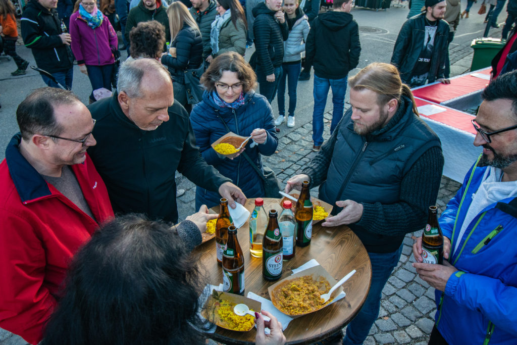 Eine Gruppe von Menschen versammelt sich um einen runden Tisch im Freien, genießt Essen von Pappschalen und hält Bierflaschen in den Händen. Die Szene ist lebhaft, andere tummeln sich im Hintergrund und tauschen Informationen über etwas aus, das wie ein Straßenfest oder Markt aussieht.