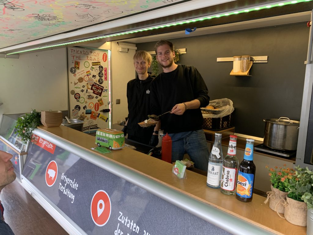 Zwei lächelnde Menschen in einem Food Truck, der Essen inmitten von bunten Aufklebern und Schriftzügen serviert. Verschiedene Gerichte, Gewürze und Getränke wie Tee und Limonade sind ausgestellt. Die Decke ist eine Leinwand mit lebendigem Dekor, die Neugier weckt und Informationen über die leckeren Angebote weckt.