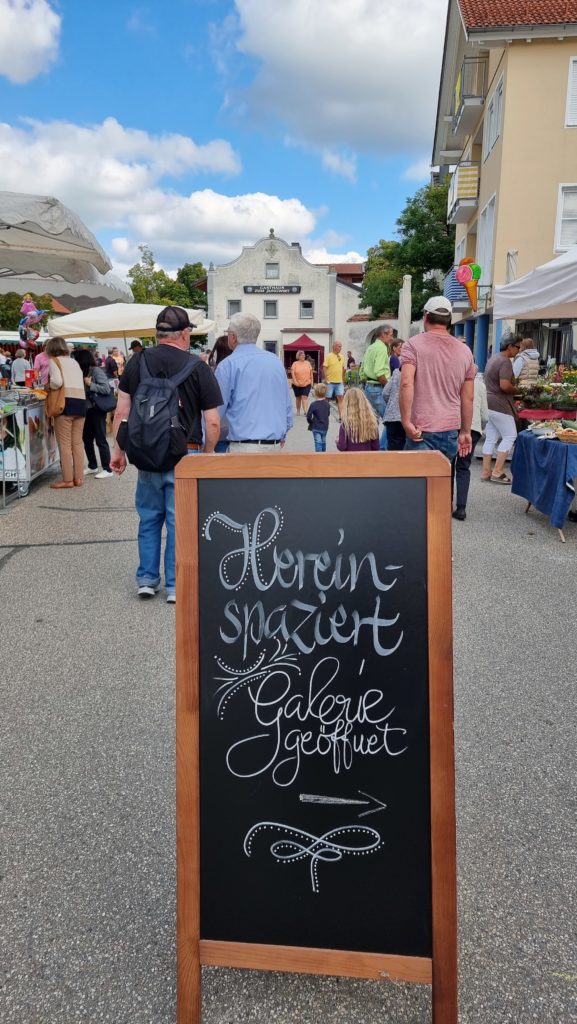 Eine Straßenmarktszene mit einem Kreideschild im Vordergrund, auf dem steht: „Herein spaziert, Galerie geöffnet.“ Informationen gibt es in Hülle und Fülle, während Menschen unter einem teilweise bewölkten Himmel zwischen den Marktständen umhergehen. Im Hintergrund sind Gebäude zu sehen.