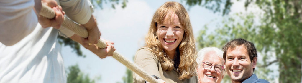 Drei Personen lächeln, während sie im Freien Tauziehen spielen. Sie halten ein Seil und die grünen Bäume im Hintergrund lassen eine Parkkulisse vermuten.