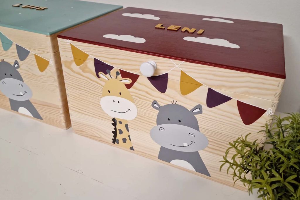 Gezeigt werden zwei Holzkisten mit Tiermotiven. Die linke Kiste hat einen grünen Deckel, auf dem ein Esel abgebildet ist, die rechte Kiste hat einen roten Deckel mit dem Namen „Leni“, darauf sind eine Giraffe und ein Nilpferd unter bunten Wimpeln zu sehen. Daneben steht eine kleine Pflanze.
