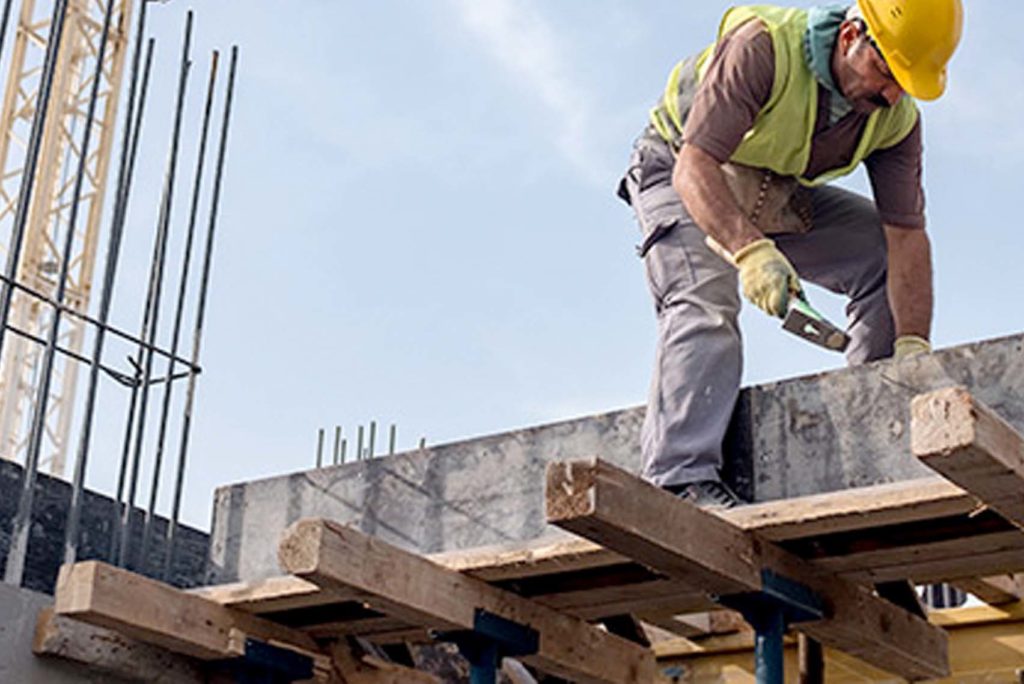 Ein Bauarbeiter mit gelbem Schutzhelm und Sicherheitsweste steht auf einer Holzplattform. Er arbeitet mit einem Handwerkzeug auf einer Betonoberfläche. Betonstahl ist sichtbar und im Hintergrund ist unter einem klaren Himmel ein Kran zu sehen.