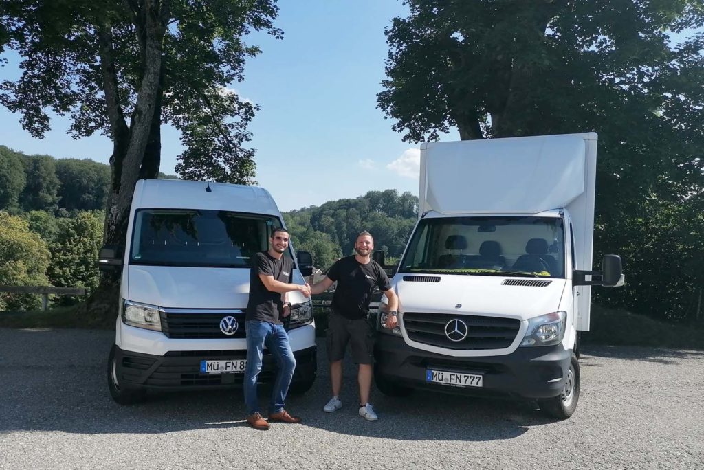 Zwei Menschen stehen lächelnd vor zwei weißen Lieferwagen, einem Volkswagen und einem Mercedes-Benz. Sie befinden sich in einer Außenkulisse mit Bäumen und einem klaren Himmel im Hintergrund.