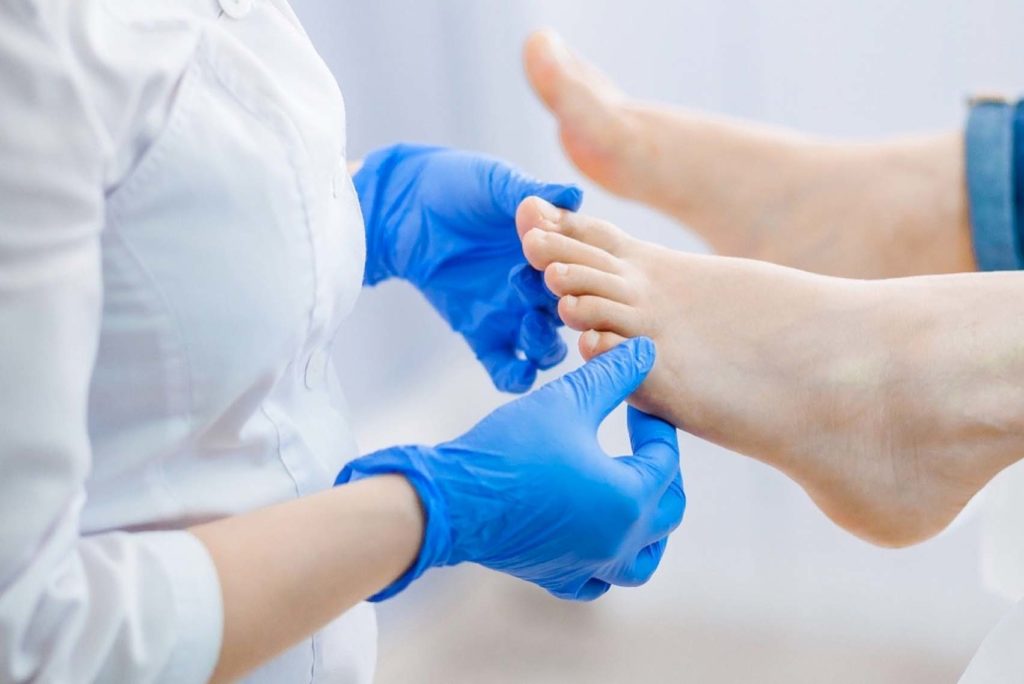 Eine Person mit blauen Handschuhen untersucht den Fuß einer anderen Person. Der Fokus liegt auf den Zehen, die der Untersuchende sorgfältig inspiziert oder behandelt. Der Hintergrund ist unscharf, wodurch die Interaktion zwischen den beiden Personen hervorgehoben wird.