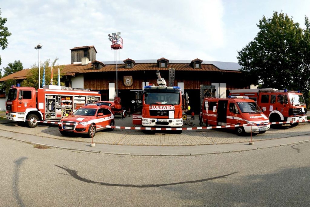 Vor einer Feuerwache mit Holzfassade parken vier Feuerwehrautos und ein roter Geländewagen. Die Fahrzeuge sind rot mit weißen Streifen und der Aufschrift „Feuerwehr“. Bäume flankieren das Gebäude, über ihm ist ein bewölkter Himmel.