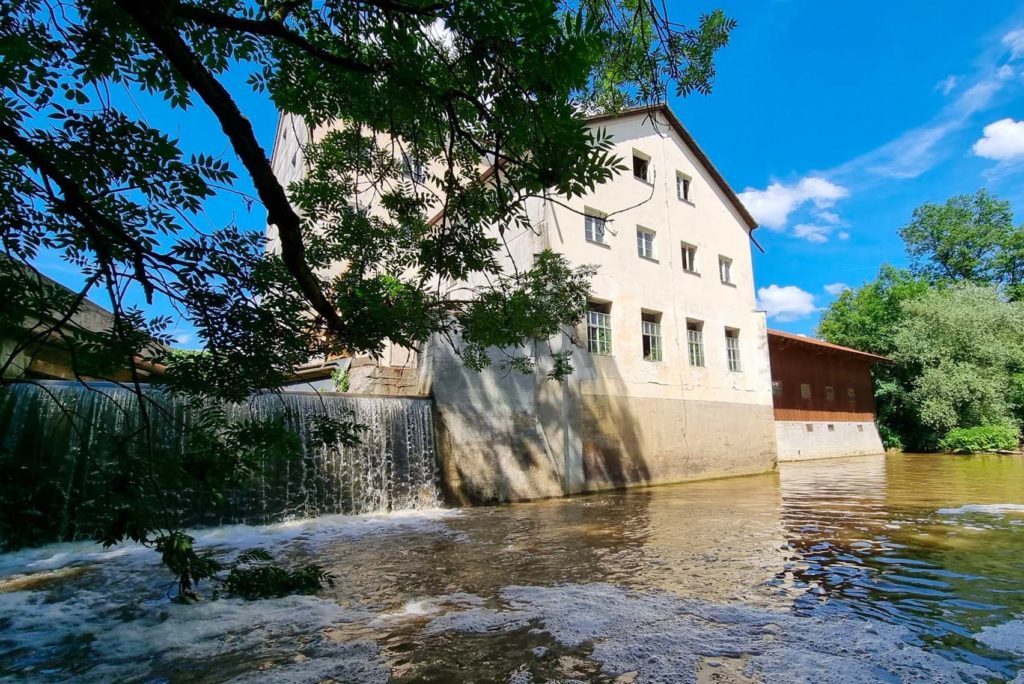 Eine ruhige Szene eines historischen Mühlengebäudes neben einem fließenden Fluss. Die Mühle hat eine rustikale Fassade mit mehreren Fenstern. Der Fluss bildet einen kleinen Wasserfall neben der Mühle, umgeben von üppigem Grün unter einem klaren blauen Himmel.