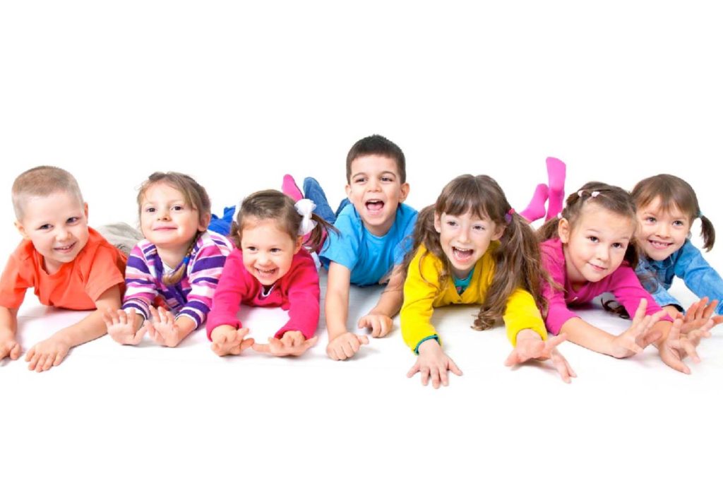 Sieben Kinder liegen auf dem Bauch, lächeln und strecken die Arme in Richtung Kamera. Sie tragen bunte Kleidung vor einem weißen Hintergrund und zeigen einen Ausdruck von Freude und Verspieltheit.