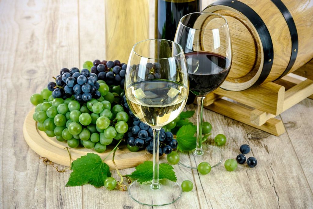Zwei Weingläser, eines mit Weißwein und eines mit Rotwein, stehen neben einem kleinen Holzfass auf einem Holztisch. Daneben steht ein Teller mit grünen und dunklen Weintrauben, auf dem einige lose Trauben verstreut liegen.