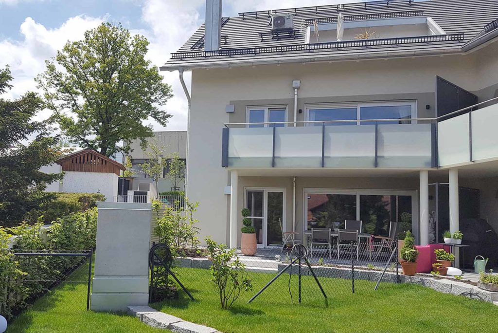 Modernes zweistöckiges Haus mit geräumigem Balkon und Terrasse. Die Terrasse ist mit Gartenmöbeln, darunter einem Tisch und Stühlen, ausgestattet. Das Haus ist von einem gepflegten Rasen und verschiedenen Pflanzen umgeben. Im Hintergrund ist unter einem teilweise bewölkten Himmel ein Baum zu sehen.