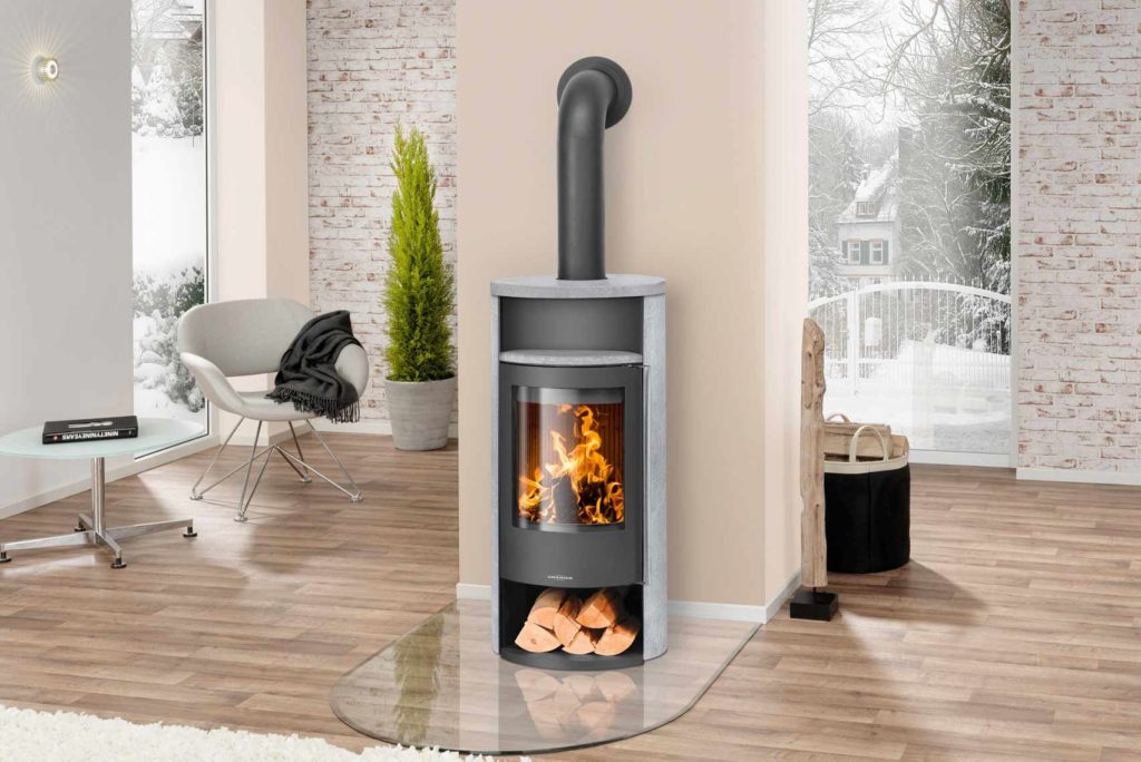 Ein modernes Wohnzimmer verfügt über einen eleganten Holzofen mit sichtbaren Flammen und darunter gelagerten Holzscheiten. Der Raum hat einen hellen Holzboden, einen weißen Sessel und große Fenster mit Blick auf eine verschneite Außenszene.