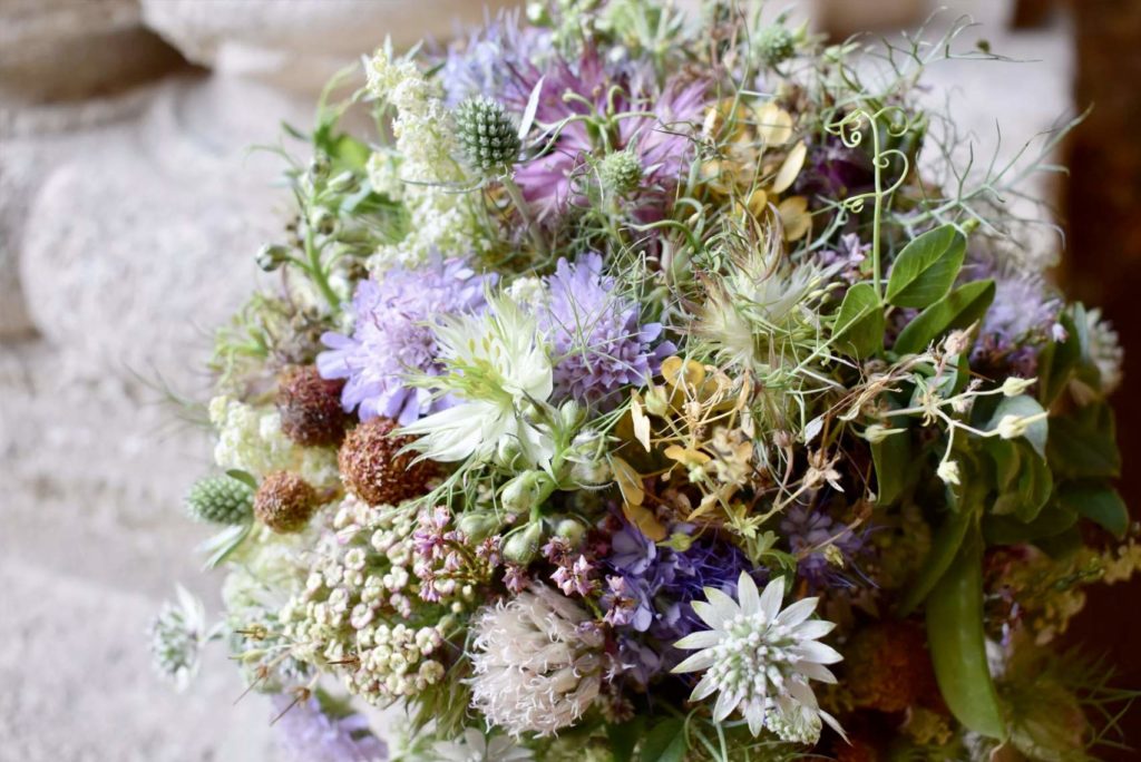 Eine Nahaufnahme eines Blumenstraußes mit einer Mischung aus Wildblumen und Grünpflanzen in verschiedenen Lila-, Weiß- und Grüntönen. Der Hintergrund ist eine weiche, verschwommene Steinoberfläche.