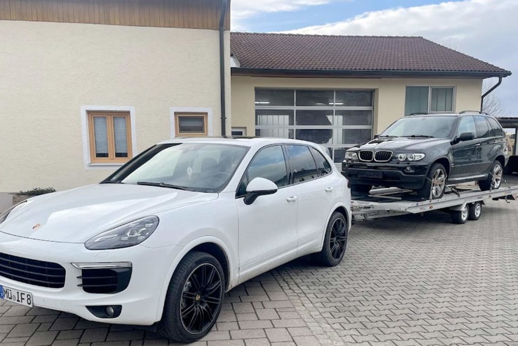 Ein weißer Porsche-SUV steht in einer Einfahrt und zieht einen Anhänger mit einem dunkelfarbigen BMW-SUV. Die Szene spielt sich vor einem cremefarbenen Haus mit einem braunen Dach und einer großen Garage ab. Der Himmel ist teilweise bewölkt.