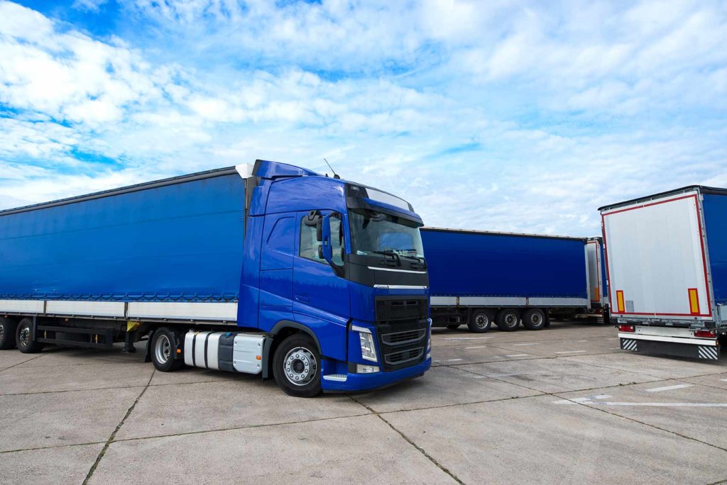 Ein blauer Sattelschlepper mit einem überdachten Anhänger steht auf einem Betonplatz. Im Hintergrund sind zwei weitere Lastwagen mit blauen und grauen Anhängern zu sehen. Der Himmel ist teilweise bewölkt.