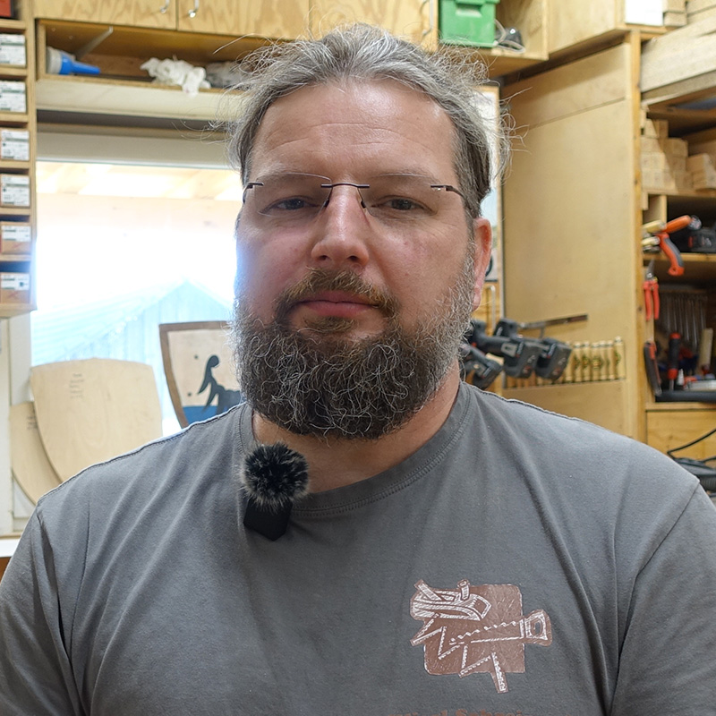 Ein Mann mit Brille und Bart steht in einer Werkstatt. Er trägt ein graues T-Shirt mit einem Aufdruck. Im Hintergrund sorgen Regale voller Werkzeuge und Materialien für eine geschäftige Atmosphäre. An seinem Hemd ist ein Mikrofon befestigt, während er über ein exklusives AmPfinger-Gutschein-Angebot spricht.