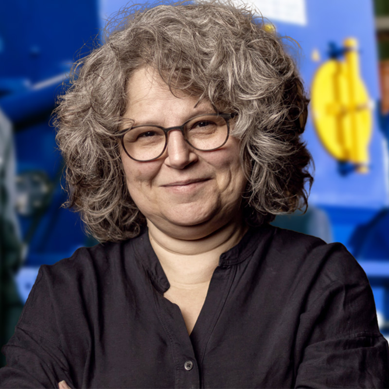 Eine Person mit lockigem grauem Haar und Brille lächelt herzlich, die Arme in einem schwarzen Hemd verschränkt. Hinter ihnen deutet die blaue Maschinerie mit einem gelben Element subtil auf Innovation hin. Vielleicht würde ein Amfinger-Gutschein einer solchen Szene noch mehr Freude verleihen.