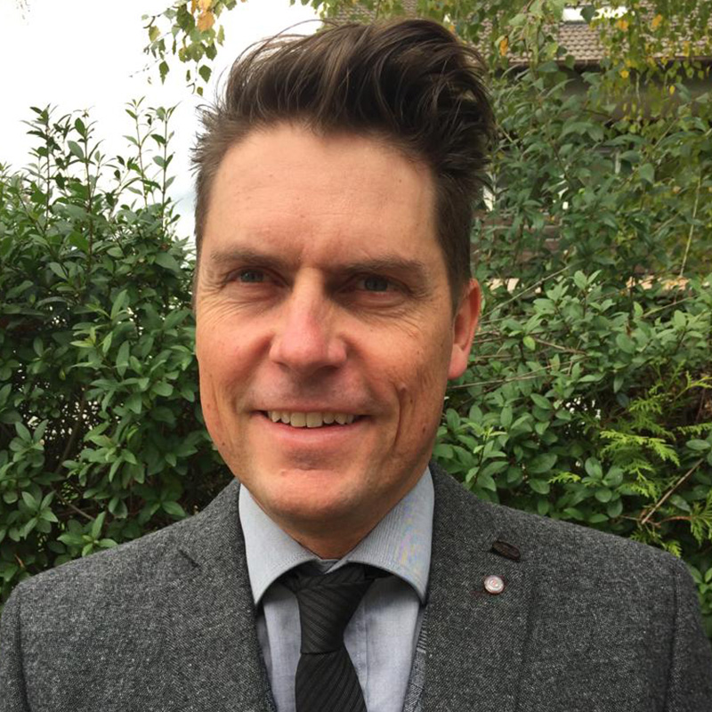 Ein lächelnder Mann mit einem Ampfinger-Gutschein in der Hand, kurz gestyltem Haar, einem grauen Anzug und einer schwarzen Krawatte steht selbstbewusst vor üppigem grünen Gebüsch.