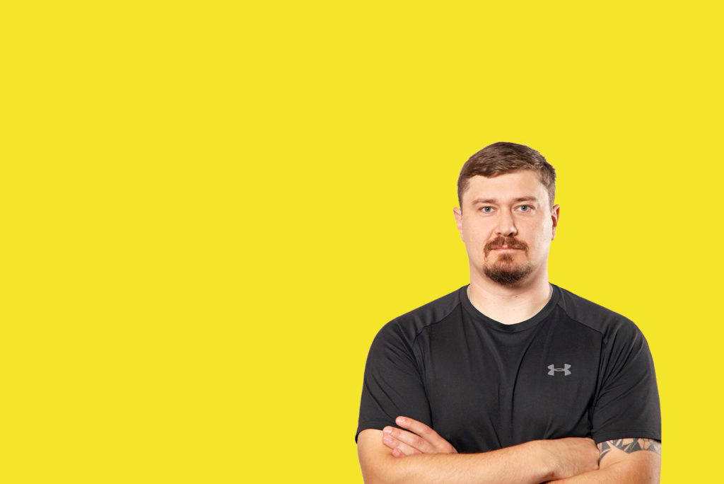 Ein Mann mit kurzen Haaren und Bart steht mit verschränkten Armen da und trägt ein schwarzes Under Armour-Shirt. Er steht vor einem leuchtend gelben Hintergrund.