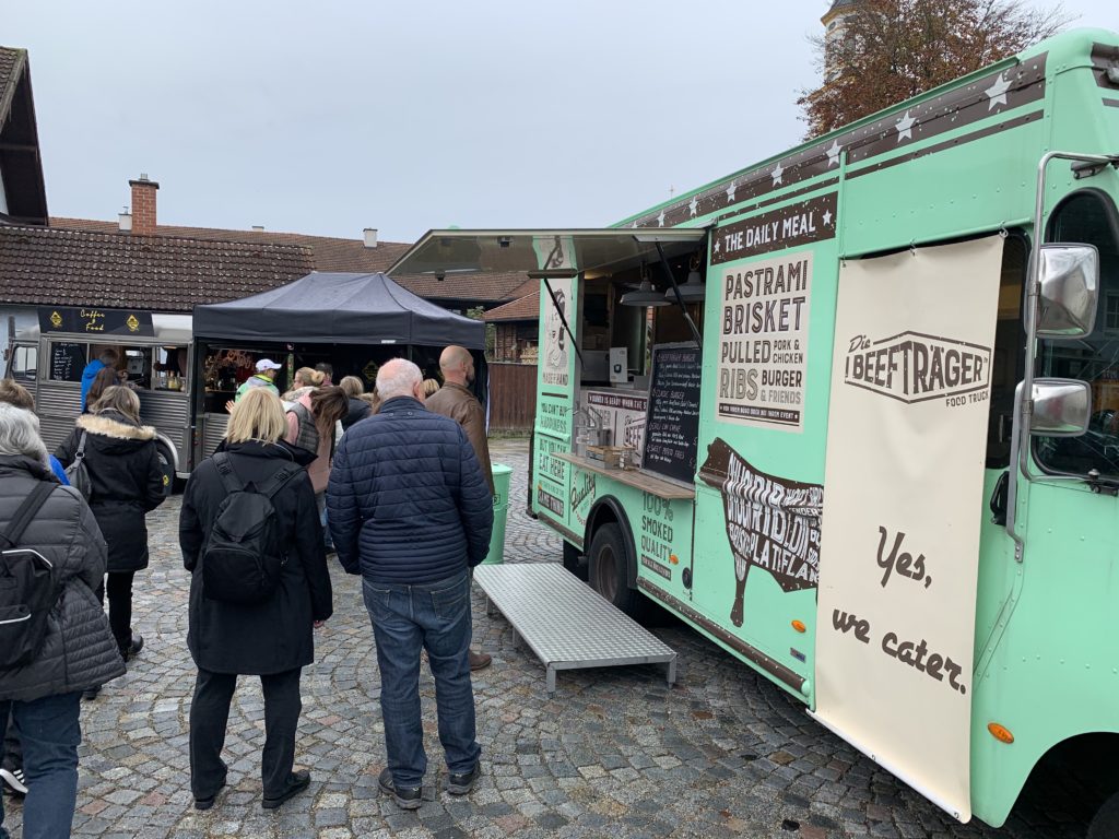 Menschen stehen Schlange vor einem grünen Imbisswagen, der Pastrami, Rinderbrust, Burger und Pommes anbietet. Am Wagen hängt ein Schild mit der Aufschrift „The Beef Trager“ und „Ja, wir liefern.“ Im Hintergrund sind ein schwarzes Zelt und Gebäude zu sehen.