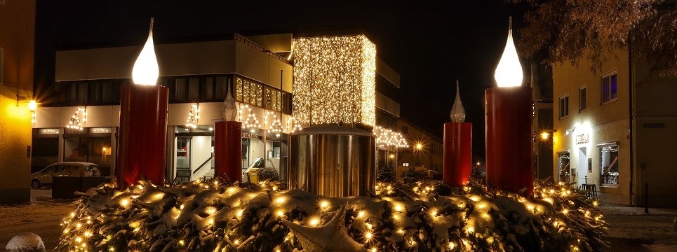Eine festliche Weihnachtsdekoration im Freien besteht aus großen roten Kerzen, die mit funkelnden gelben Lichtern geschmückt sind und von einer üppigen, beleuchteten Girlande umgeben sind. Die Szene spielt vor dem Hintergrund sanft beleuchteter Gebäude bei Nacht.