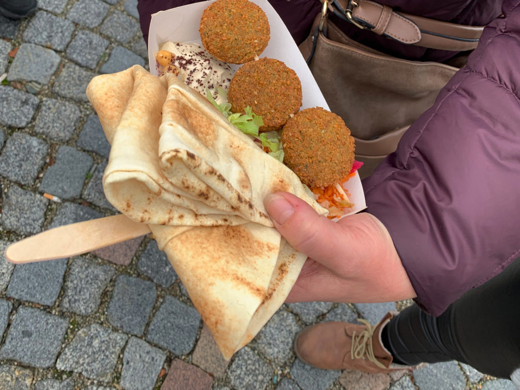 Eine Person in einer kastanienbraunen Jacke steht auf einem Kopfsteinpflaster und genießt ein mit Falafel, Salat und Belag gefülltes Pita-Brot. Das hübsch verpackte Pita-Brot mit einem Holzbesteck darin könnte der perfekte Leckerbissen sein, den man mit einem Ampfinger-Gutschein einlösen kann.