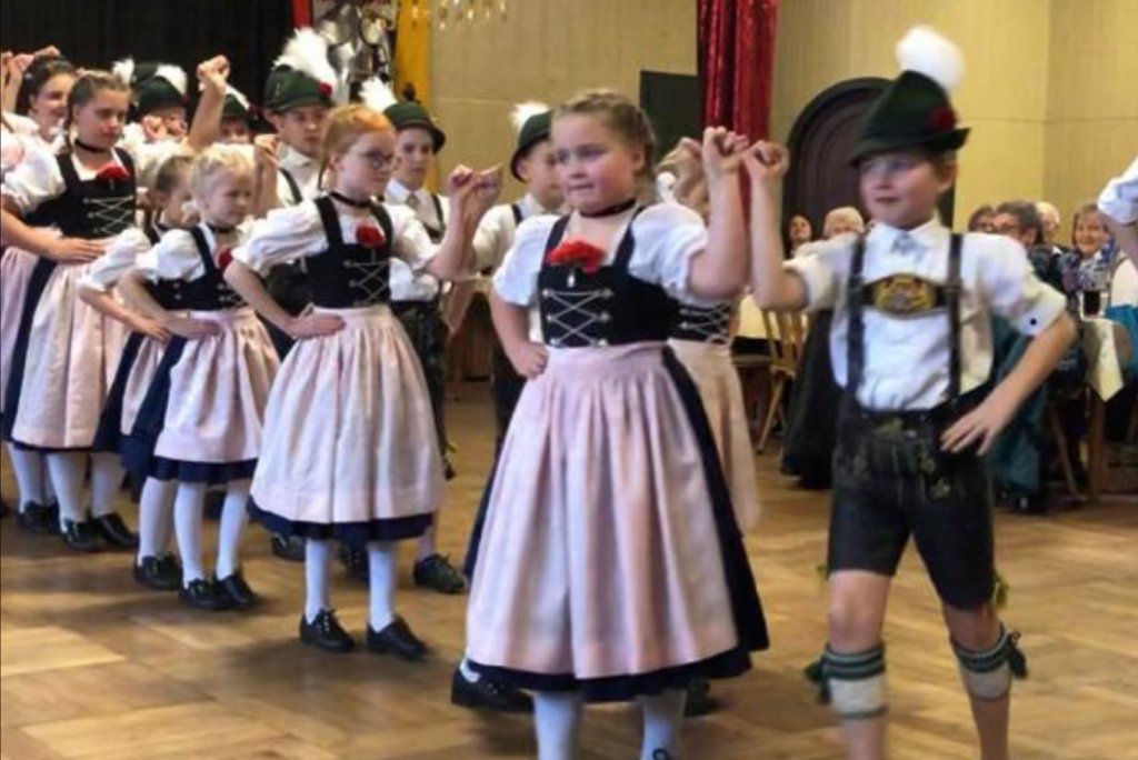 Kinder in traditioneller bayerischer Kleidung tanzen in einem Raum. Die Mädchen tragen Dirndl mit rosa Schürzen und die Jungen Lederhosen mit grünen Hüten. Sie halten sich an den Händen und führen einen Volkstanz auf, während im Hintergrund ein Publikum zusieht.