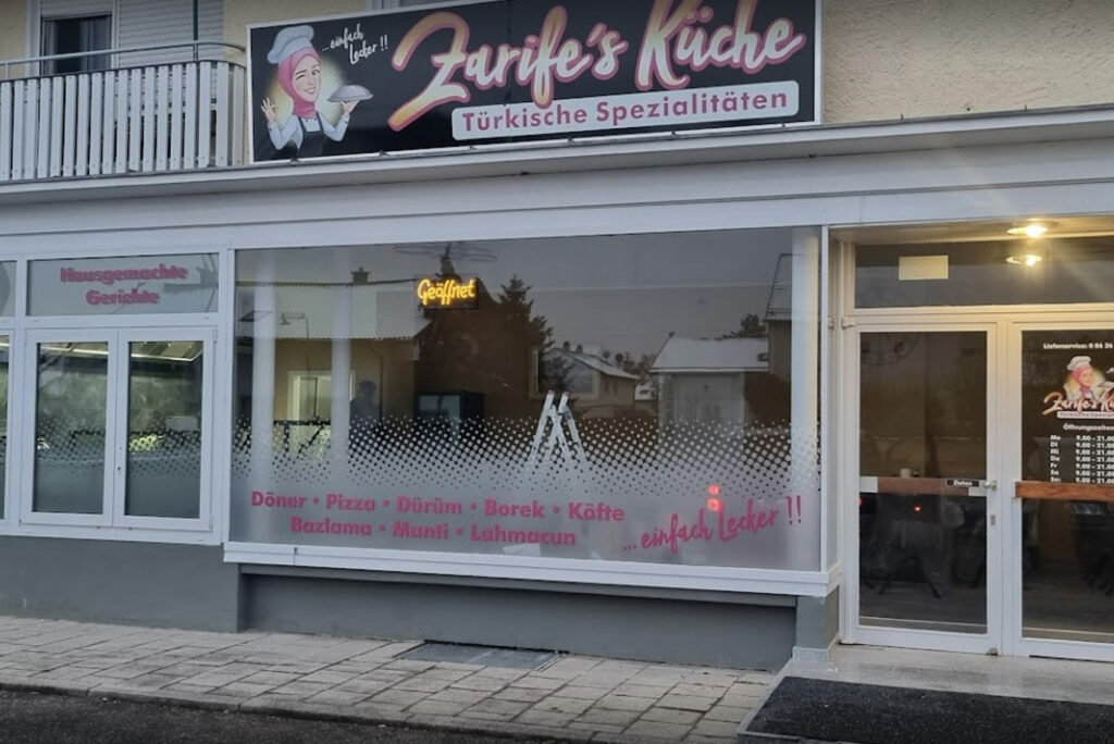 Straßenansicht eines türkischen Spezialitätenrestaurants mit einem Schild „Zarife’s Küche“ und Bildern von Gerichten wie Döner, Pizza und Börek im Fenster. Das Gebäude ist hell und hat einen Balkon darüber. Im Schaufenster ist eine Leiter zu sehen.