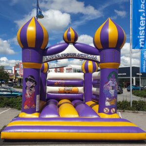 Ein farbenfrohes aufblasbares Hüpfburg-Design mit violetten und gelben Türmen. Die Struktur trägt die Aufschrift „Super Traum Schloss“ und an den Seiten sind Comicfiguren abgebildet. Sie steht an einem sonnigen Tag im Freien in der Nähe von Geschäften.