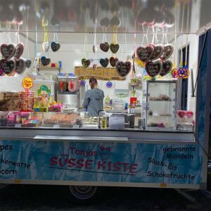 Eine Frau steht in „Tanjas Süße Kiste“, einem farbenfrohen Imbissstand, der Süßigkeiten anbietet. Vorne hängen herzförmige Dekorationen und verschiedene Bonbons, Slushies und Konfekt sind ausgestellt. Das Schild ist auf Deutsch, was auf ein verspieltes Thema hindeutet.