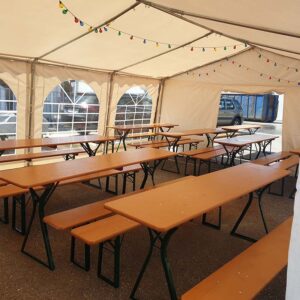 Innenansicht eines großen, seitlich offenen Zelts mit in Reihen angeordneten hölzernen Picknicktischen und Bänken. Von der Decke hängen bunte Lichterketten. Durch die gewölbten Fenster fällt helles Tageslicht.