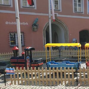 Vor einem Gebäude mit der Aufschrift "Stadtbibliothek" ist eine kleine Kindereisenbahn mit einer Lokomotive und zwei bunten Waggons aufgebaut. Sie ist von einem Holzzaun umgeben, auf einem kleinen Schild steht "AUSGANG".