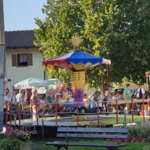 Ein buntes Karussell mit sonnenförmiger Spitze steht auf einem lebhaften Jahrmarkt im Freien. Menschen haben sich um das Karussell versammelt, einige sitzen, im Hintergrund sind ein grünes Zelt und ein Haus zu sehen. Im Vordergrund sind eine Bank und Blumen zu sehen.
