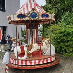 Ein kleines, altes Karussell mit zwei weißen Pferden und roten Sitzen steht im Freien. Das rot-weiß gestreifte Dach ist mit dekorativen Paneelen versehen. Im Hintergrund laufen zwei Menschen in Jacken in der Nähe von Grünflächen und einem Gebäude.