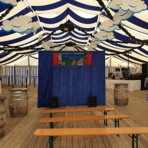 Ein mit blauen und weißen Streifen dekoriertes Zelt, in dem sich ein Puppentheater mit blauem Vorhang und kleiner Bühne befindet. Auf dem Holzboden sind Holzbänke und Fässer als Sitzgelegenheiten aufgestellt. Von der Decke hängen Wolkendekorationen.