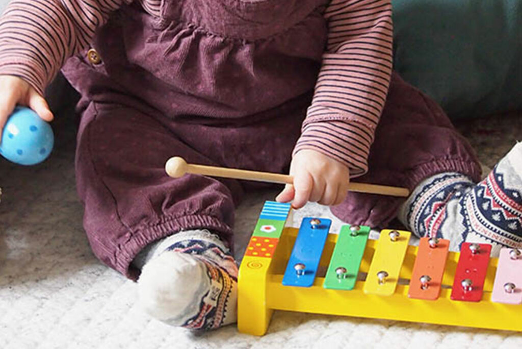 Ein Kind in gemütlicher Kleidung spielt mit einem bunten Xylophon. In der einen Hand hält es einen Schlägel, in der anderen einen blauen eiförmigen Shaker. Das Xylophon hat bunt bemalte Tasten und liegt auf einer weichen Oberfläche.