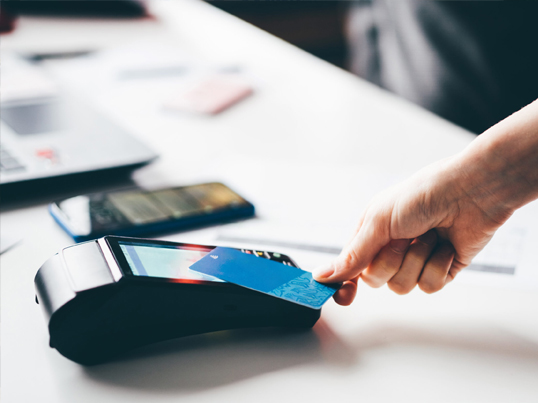 Eine Person hält eine blaue Kreditkarte über ein Kartenlesegerät zum kontaktlosen Bezahlen. Daneben liegen ein Smartphone und ein Laptop auf einer weißen Fläche. Die Szene vermittelt eine moderne, digitale Transaktionsumgebung.