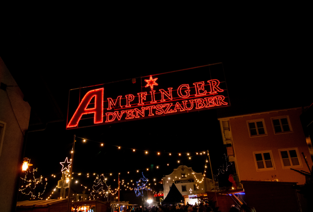 Eine festliche Nachtszene auf einem Weihnachtsmarkt mit einem leuchtend roten Schild mit der Aufschrift „Ampfinger Adventszauber“. Der Bereich ist mit Lichterketten und dekorierten Ständen geschmückt. Eine Menschenmenge genießt die lebhafte Atmosphäre.