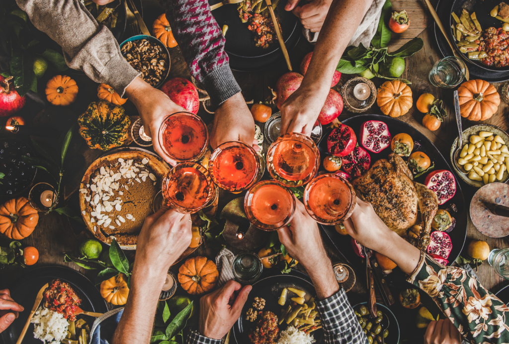 Eine Gruppe von Menschen stößt an einem festlich gedeckten Tisch mit Gläsern eines rosigen Getränks an. Der Tisch ist mit herbstlichen Speisen geschmückt, darunter Granatäpfel, Mini-Kürbisse und verschiedene Gerichte. Die Szene strahlt eine warme, festliche Atmosphäre aus.