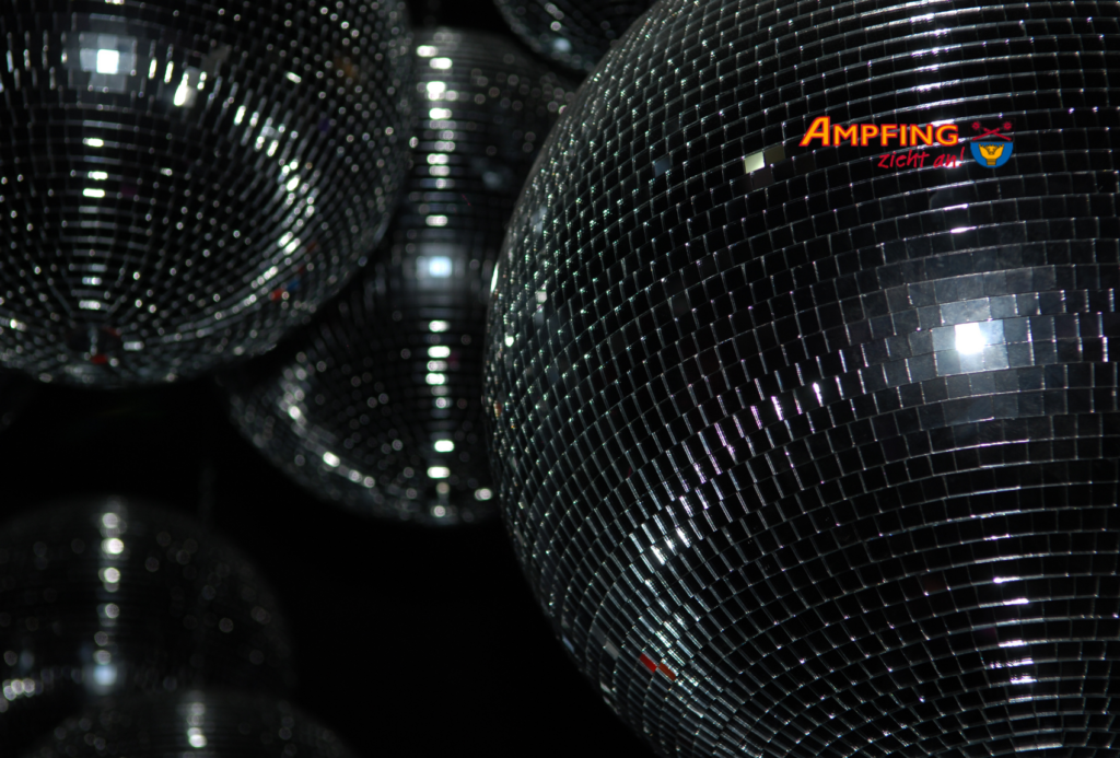 Nahaufnahme mehrerer glänzender Discokugeln, die das Licht in einem dunklen Raum reflektieren, mit einem Logo mit der Aufschrift "Ampfing zeigt sich" und einem kleinen blau-gelben Emblem in der oberen rechten Ecke.