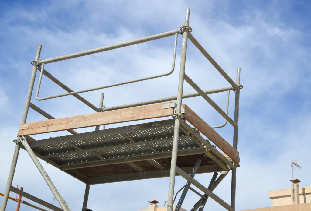 Ein Gerüst aus Metall und Holz vor einem blauen Himmel mit dünnen Wolken. Die Konstruktion wirkt stabil und ist Teil einer Baustelle, die durch das klare, helle Tageslicht hervorgehoben wird.