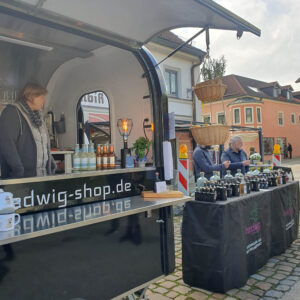 Ein schwarzer Foodtruck mit der Aufschrift "hedwig-shop.de" steht auf einer Kopfsteinpflasterstraße. Draußen stehen Flaschen auf Tischen, und drei Personen stehen hinter den Tischen und unterhalten sich mit Passanten.