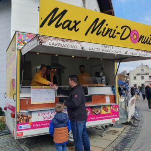 Ein Mann und ein Kind stehen vor einem gelben Donut-Imbisswagen mit der Aufschrift "Max' Mini-Donuts" und unterhalten sich mit dem Personal im Inneren. Auf der Speisekarte des Wagens sind verschiedene Donut-Varianten und Heißgetränke abgebildet, im Hintergrund sind Häuser und Menschen zu sehen.