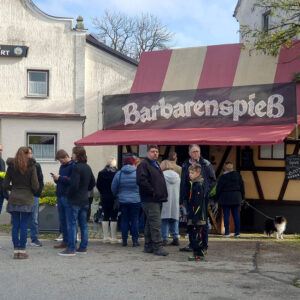 Eine Gruppe von Menschen steht in einer Schlange vor einem Imbissstand mit einer rot-weiß gestreiften Markise und einem Schild mit der Aufschrift "Barbarenspieß". Einige Personen unterhalten sich, während andere in die Kamera schauen. Auf der rechten Seite ist ein Hund zu sehen.